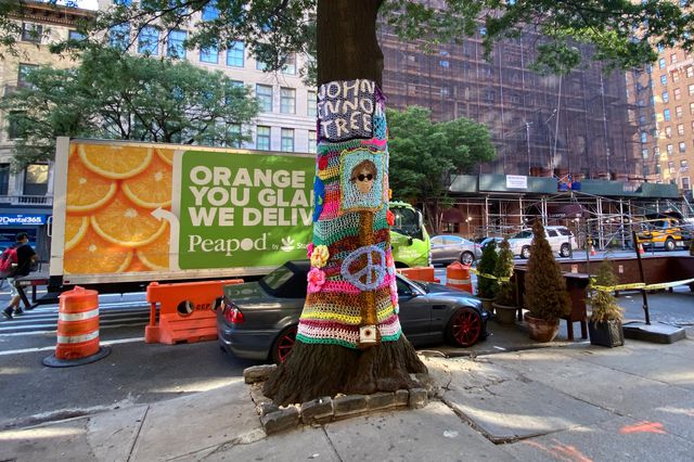 A tree on West 79th Street with a knit artwork on it, with the words "John Lennon Tree" and an likeness of Lennon, plus a peace sign and flowers